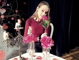 Młoda dziewczyna w bordowej bluzce siedzi przy stole w ręce trzyma różę. Na stole stoi świecznik. 